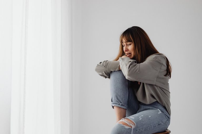 A woman in jeans sits against a white curtain, expressing emotion.